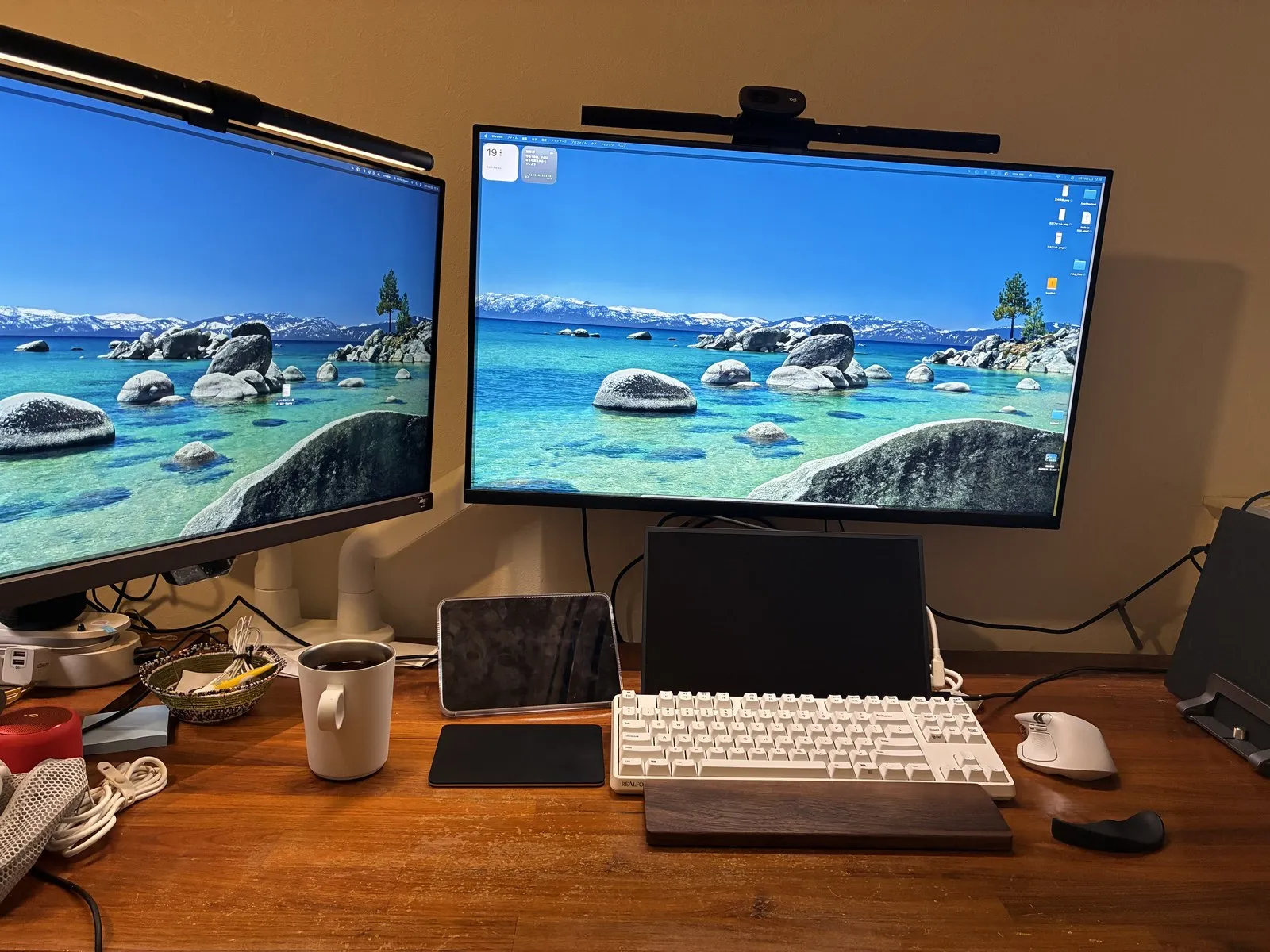 木製天板のデスク全景。白いアームでサイズの違うモニター2枚を並べ、手前に白いREALFORCEキーボードと木製のパームレスト、右にMX Masterマウスが置かれている