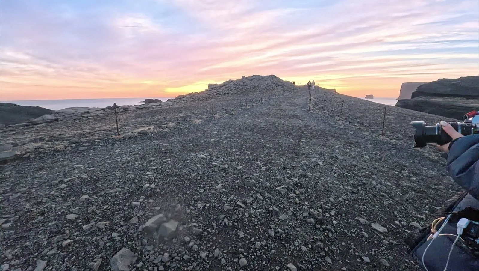 夕暮れの岬。砂利の丘の先に海と岩が見え、右手にカメラを持った手が入っている
