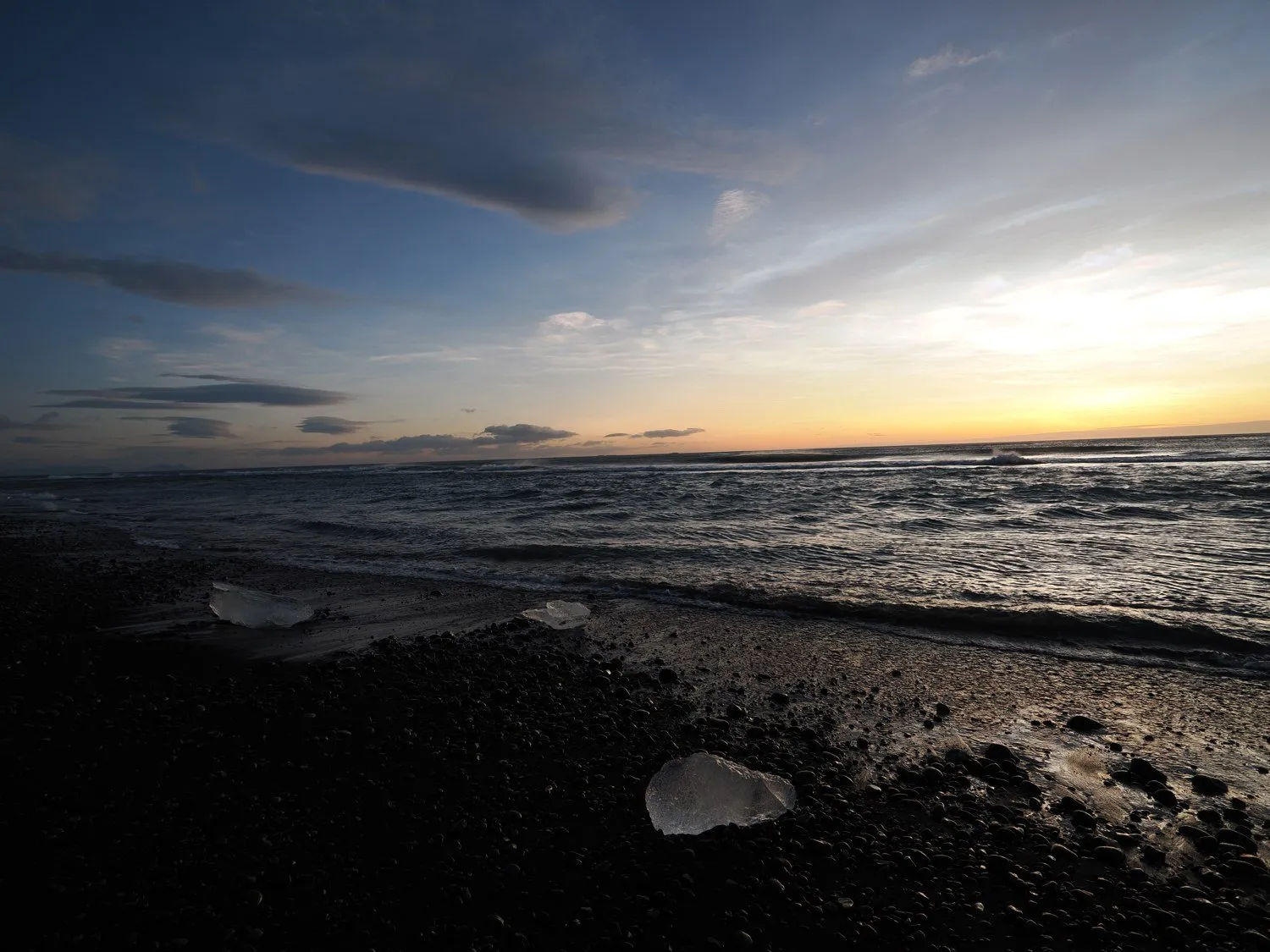 黒い砂浜に透明な氷の塊が点在し、奥に朝焼けの空と波