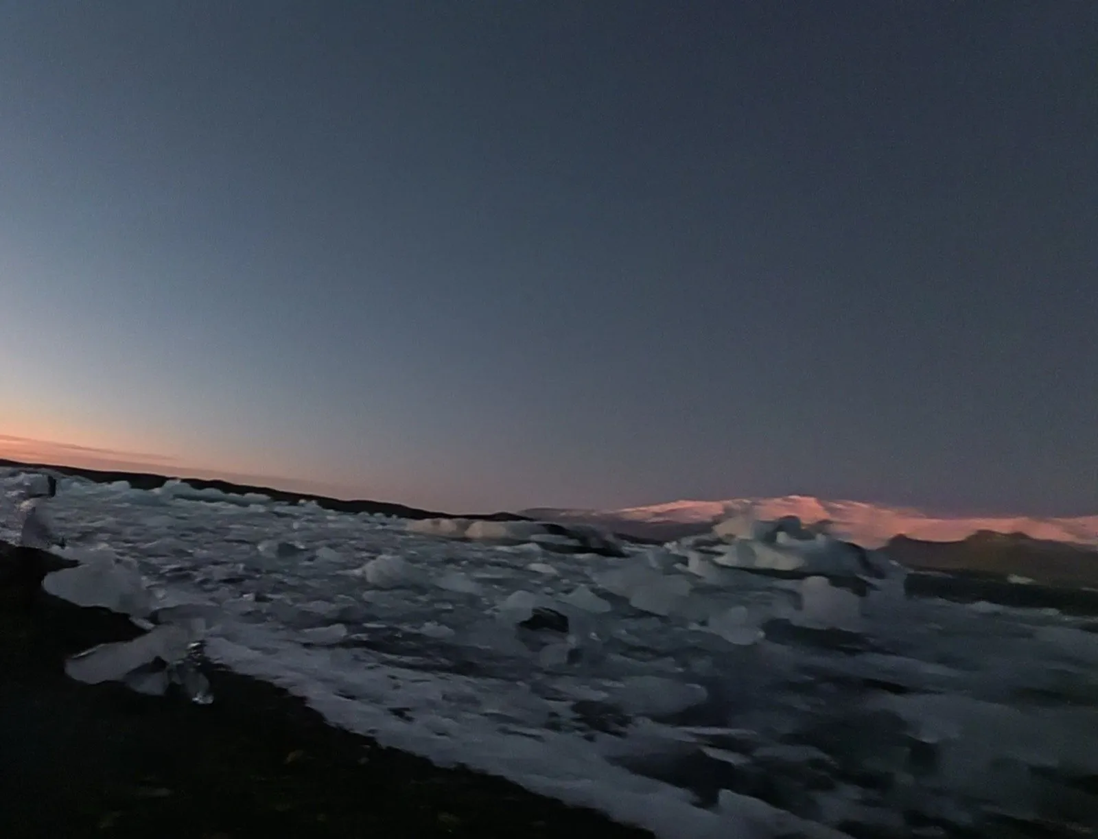 氷河湖の岸に打ち上げられた氷の塊と、遠くに夕日を受けた雪山